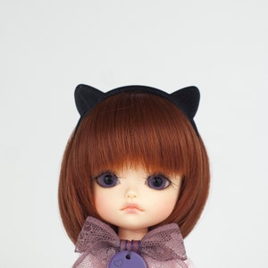May include: A doll with brown hair and a black cat ear headband. The doll has light yellow skin and blue eyes. The doll is wearing a purple and white bow.