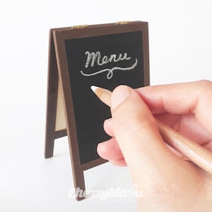 May include: A small wooden chalkboard easel with the word "Menu" written in white chalk on the black surface. A hand is holding a white chalk pencil and writing on the chalkboard.