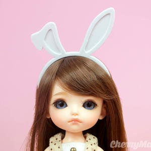 Può includere: Una bambola con lunghi capelli castani che indossa una fascia per capelli con orecchie di coniglio bianche. La bambola ha gli occhi azzurri e una carnagione giallo chiaro. La bambola indossa un vestito a pois marroni e bianchi.