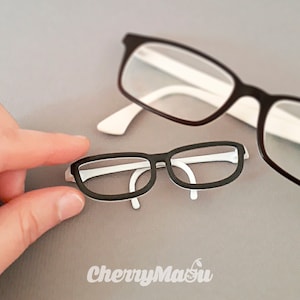 Peut inclure: Deux paires de lunettes, une noire avec des branches blanches et une plus petite noire avec des branches blanches. La plus petite paire est tenue par une main.
