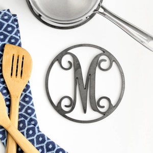 May include: A blue and white patterned kitchen towel with two wooden spoons and a silver pot on a black trivet. The trivet has a metal cutout of the letter M.