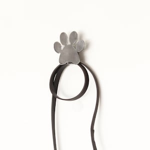 Puede incluir: Un gancho de pared de metal con forma de huella de pata que sostiene una correa de cuero marrón oscuro para perros. El gancho está montado en una pared blanca. La correa tiene un cierre de metal.