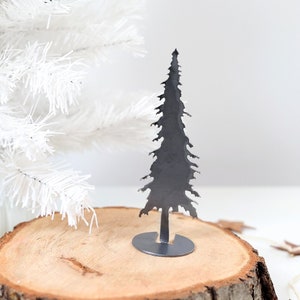 May include: A metal silhouette of a Christmas tree on a wooden slice with a white artificial Christmas tree in the background. A garland of brown leather stars is on the table.