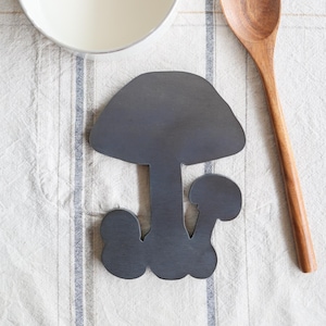 Puede incluir: Una cacerola blanca con asa dorada, una cuchara de madera y un salvamanteles en forma de seta gris oscuro descansan sobre un mantel a rayas blanquecino. La cacerola contiene un líquido blanco.