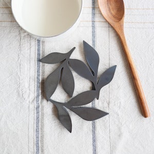 May include: A set of black metal leaf-shaped trivets on a white and brown striped tablecloth, next to a white enamel saucepan with a gold handle and a wooden spoon.