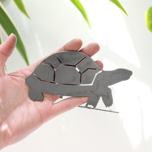 K&ouml;nnte beinhalten: Eine Metallsilhouette einer Schildkr&ouml;te, aus Blech ausgeschnitten, in einer Hand. Die Schildkr&ouml;te ist nach rechts gerichtet und hat einen detaillierten Panzer.