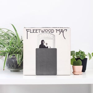 May include: A white vinyl record with the text "Fleetwood Mac" on the cover. The record is standing upright on a white shelf with a gray bookend. There are two small green plants in pots on the shelf.