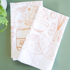 May include: Two white kitchen towels with a brown outline of baking supplies. The towels feature illustrations of a mixing bowl, measuring cups, a whisk, a loaf of bread, a jar of vanilla extract, a jar of baking powder, a jar of flour, and a rolling pin.