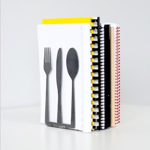 May include: A silver metal bookend shaped like a fork, knife, and spoon. The bookend is holding up three spiral bound notebooks with yellow, black, and brown covers.