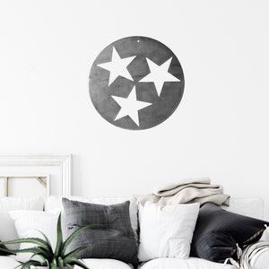 Puede incluir: Una pieza de arte mural circular de metal con un acabado gris oscuro y tres estrellas blancas recortadas en formaci&oacute;n triangular. El dise&ntilde;o est&aacute; inspirado en la bandera del estado de Tennessee, mostrando una est&eacute;tica industrial limpia.