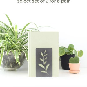 May include: A pair of metal bookends with a leafy design. The bookends are gray and have a cutout design of a branch with leaves. The bookends are shown holding a book on a white shelf. The text "bookends are sold individually, select set of 2 for a pair" is displayed above the bookends.
