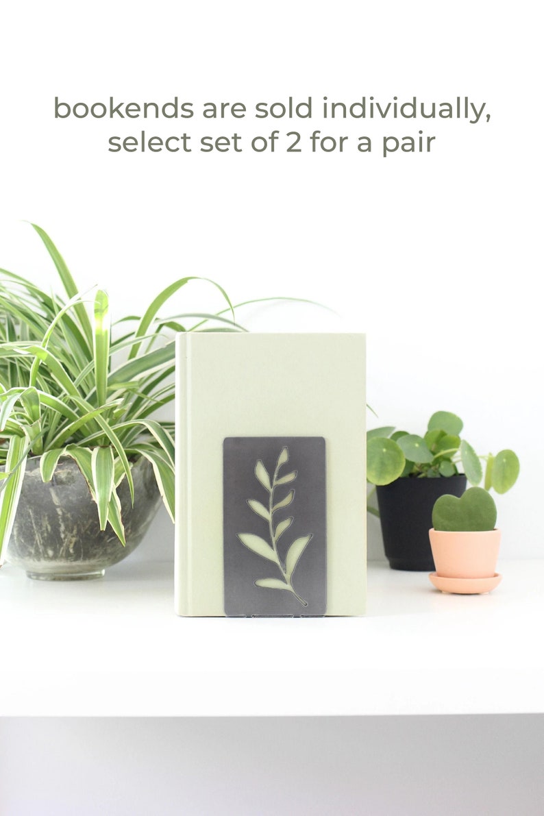 May include: A pair of metal bookends with a leafy design. The bookends are gray and have a cutout design of a branch with leaves. The bookends are shown holding a book on a white shelf. The text "bookends are sold individually, select set of 2 for a pair" is displayed above the bookends.