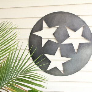 Puede incluir: Una pieza de arte mural circular de metal gris oscuro con tres formas de estrellas recortadas dispuestas en un patr&oacute;n triangular. La superficie del metal tiene un acabado mate y liso con sutiles variaciones de tono, creando un aspecto industrial.