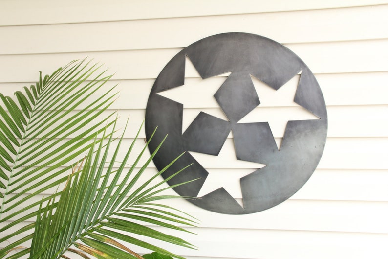Puede incluir: Una pieza de arte mural circular de metal gris oscuro con tres formas de estrellas recortadas dispuestas en un patr&oacute;n triangular. La superficie del metal tiene un acabado mate y liso con sutiles variaciones de tono, creando un aspecto industrial.