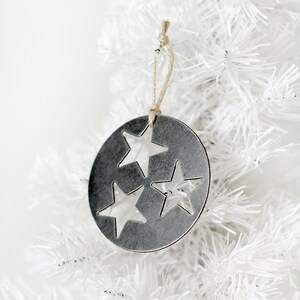May include: A silver metal Christmas ornament shaped like a circle with three cut-out stars. The ornament is hanging from a white artificial Christmas tree.