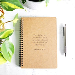 May include: A spiral-bound notebook with a tan, textured cover featuring the printed text: The righteous man walks with integrity; blessed are his children after him. Proverbs 20:7. A silver metal pen is positioned to the right of the notebook.