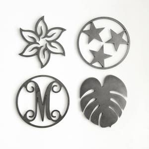 May include: Four metal coasters with different designs. The first coaster features a flower design, the second coaster features three stars, the third coaster features the letter M, and the fourth coaster features a leaf design.