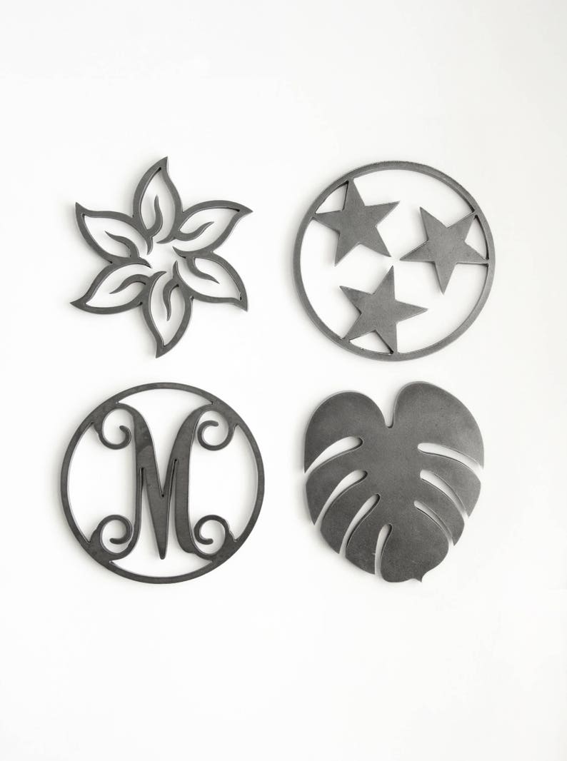 May include: Four metal coasters with different designs. The first coaster features a flower design, the second coaster features three stars, the third coaster features the letter M, and the fourth coaster features a leaf design.