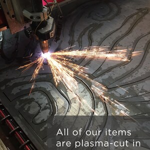 Puede incluir: Un cabezal de m&aacute;quina de corte por plasma emite chispas de color naranja brillante mientras corta un patr&oacute;n de remolinos en una l&aacute;mina de metal gris oscuro. El texto dice: All of our items are plasma-cut in Tennessee, USA. La maquinaria tiene componentes rojos y plateados.