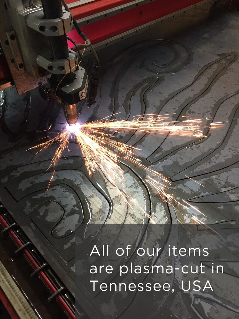 Puede incluir: Un cabezal de m&aacute;quina de corte por plasma emite chispas de color naranja brillante mientras corta un patr&oacute;n de remolinos en una l&aacute;mina de metal gris oscuro. El texto dice: All of our items are plasma-cut in Tennessee, USA. La maquinaria tiene componentes rojos y plateados.