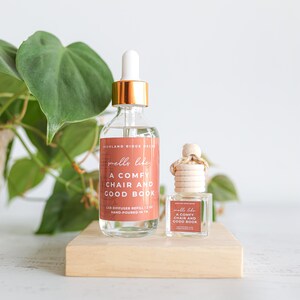 May include: Two glass bottles with the text "smells like A COMFY CHAIR AND GOOD BOOK" on a wooden block. One bottle is a car diffuser refill with a dropper, the other is a car air freshener. A green plant is in the background.