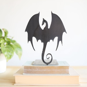 May include: Black metal dragon silhouette sculpture with wings spread, standing on a stack of two books. The dragon has a curled tail and a detailed head.