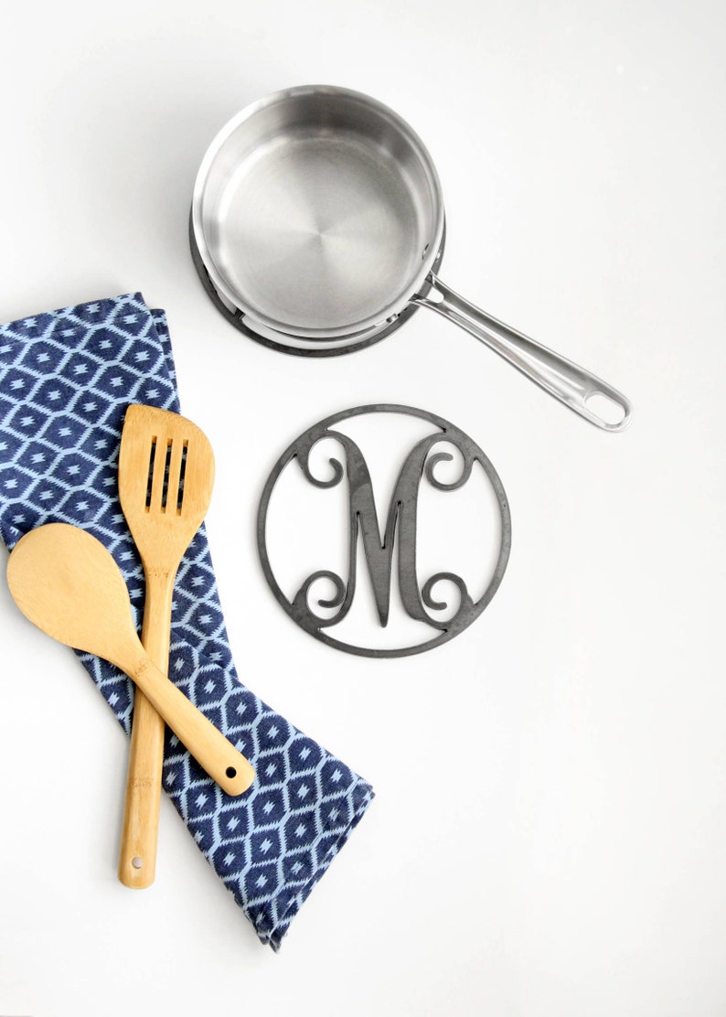 May include: A blue and white patterned kitchen towel with two wooden spoons and a silver pot on a black trivet. The trivet has a metal cutout of the letter M.