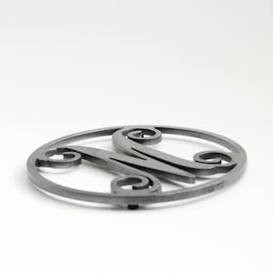 May include: A silver metal trivet with a decorative letter M in the center. The trivet is round and has a smooth surface.