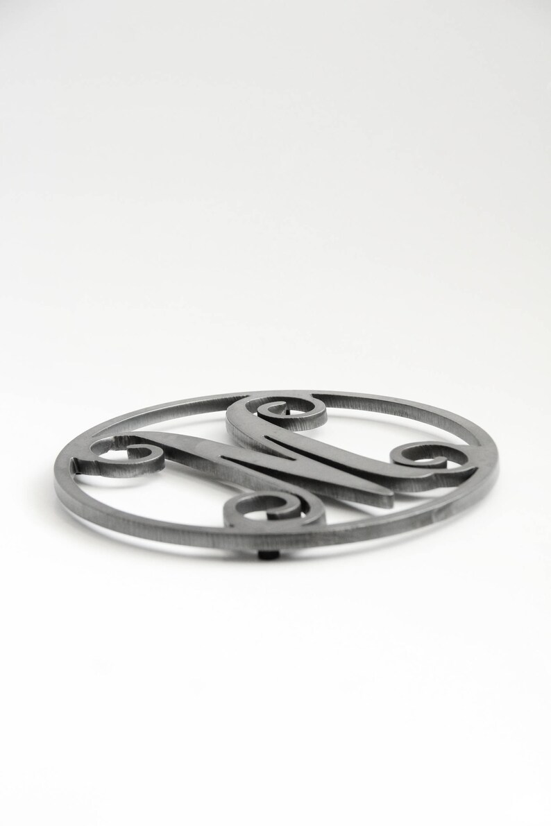May include: A silver metal trivet with a decorative letter M in the center. The trivet is round and has a smooth surface.