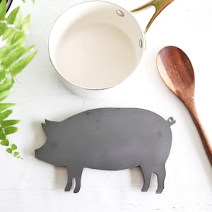 May include: A top-down shot of kitchen items on a white surface. A gray pig-shaped trivet is in the center, with a wooden spoon and a small saucepan with a gold handle. Green fern leaves are in the background.