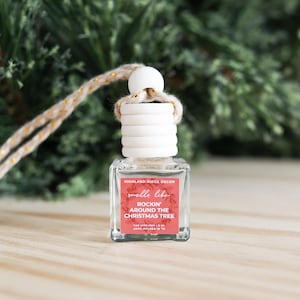 May include: A clear glass car air freshener with a white wooden top and a decorative string. The label reads "smells like Rockin' Around the Christmas Tree" in red text. The background is a blurred Christmas tree.
