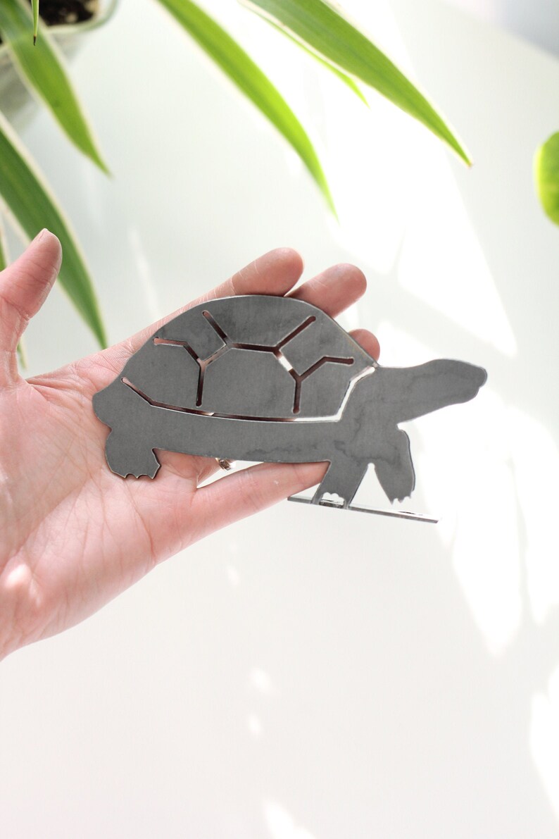 K&ouml;nnte beinhalten: Eine Metallsilhouette einer Schildkr&ouml;te, aus Blech ausgeschnitten, in einer Hand. Die Schildkr&ouml;te ist nach rechts gerichtet und hat einen detaillierten Panzer.
