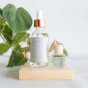 May include: A clear glass bottle with a gold-colored top and a white dropper, next to a small square car air freshener. Both are labeled "Walking in a Garden After the Rain." The products sit on a wooden block with green foliage in the background.
