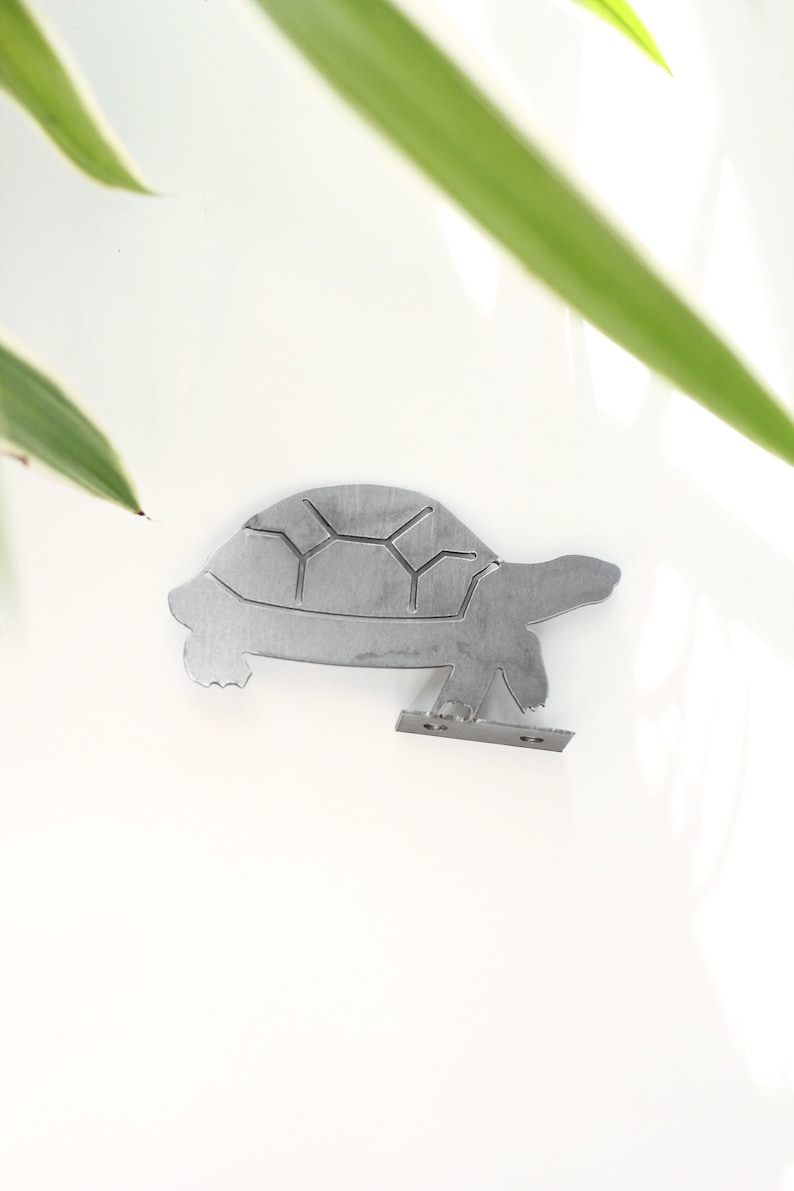 K&ouml;nnte beinhalten: Ein silberner Wandhaken in Form einer Schildkr&ouml;te aus Metall. Die Schildkr&ouml;te befindet sich in einer Gehposition, wobei Kopf und Beine sichtbar sind. Der Haken ist an der Unterseite der Schildkr&ouml;te befestigt.