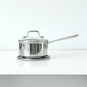 May include: A silver saucepan with a glass lid and a handle sits on a gray heat-resistant mat. The saucepan is on a white countertop.