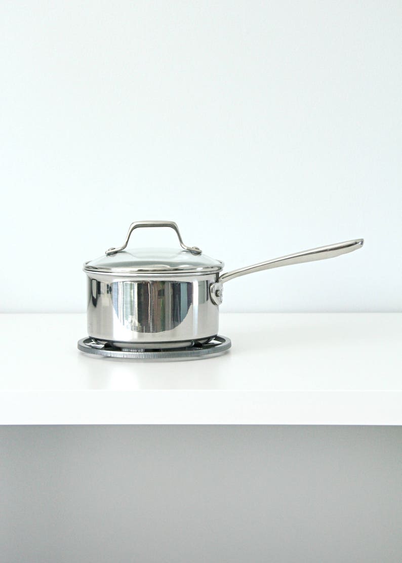 May include: A silver saucepan with a glass lid and a handle sits on a gray heat-resistant mat. The saucepan is on a white countertop.
