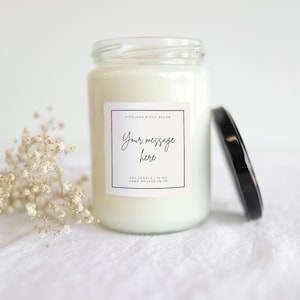 May include: A white soy candle in a clear glass jar with a black lid. The label reads "Your message here" in script font. The candle is 10 oz and hand-poured in TN. A sprig of white flowers is next to the candle.