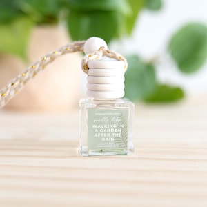May include: A small, clear glass car air freshener with a wooden cap and a braided cord. The label reads "Smells like Walking in a Garden After the Rain." The background includes green foliage and a light-colored wooden surface.