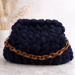 May include: A navy blue, chunky knit clutch bag with a tortoiseshell chain strap. The bag has a rounded shape and a soft, textured appearance. The chain is a warm, amber color.