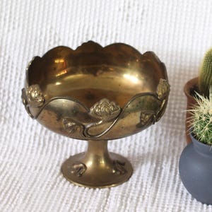 Pode incluir: Uma taça de latão com pedestal, borda recortada e detalhes florais em relevo, acompanhada por três pequenos cactos em dois vasos arredondados cinza-escuro fosco e um vaso cilíndrico marrom sobre uma superfície de tecido branco.