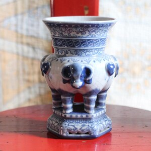 Op de afbeelding: Een blauw-witte keramische vaas met een uitlopende rand, ingewikkelde geometrische patronen en driedimensionale ramshoofdmotieven. Het object rust op een zeshoekige voet met opengewerkt detail en heeft een glanzende, craquelé glazuurafwerking.