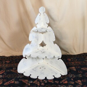 May include: A three-tiered corner shelf shaped like a stylized Christmas tree. The unit is constructed from white painted wood with a distressed, weathered finish showing dark undertones. It features three triangular shelves of varying sizes.