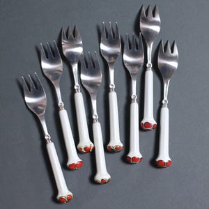 May include: Nine silver forks with white handles and red and gold accents at the base. The forks are arranged on a gray surface. The tines of the forks are visible, and the handles are cylindrical.