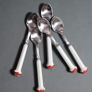 Puede incluir: Seis cucharas plateadas con mangos blancos y detalles rojos. Las cucharas están dispuestas sobre una superficie gris. Los mangos tienen forma bulbosa con un diseño floral rojo en la base.