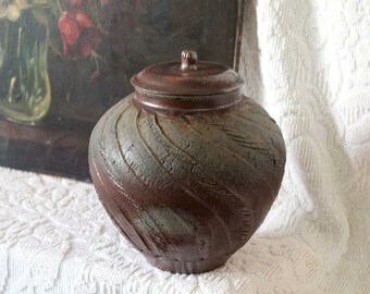 Op de afbeelding: Een bruine keramische pot met een deksel, met een swirl-ontwerp en een getextureerd oppervlak. De pot staat op een wit stoffen oppervlak.