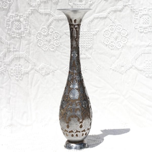 Könnte beinhalten: Eine hohe, silberfarbene Vase mit einem ausgestellten oberen Rand und aufwendigen floralen Mustern. Die Vase hat einen runden Sockel und eine längliche Form. Das Design ist symmetrisch, und die Vase wirft einen Schatten auf die weiße Oberfläche.