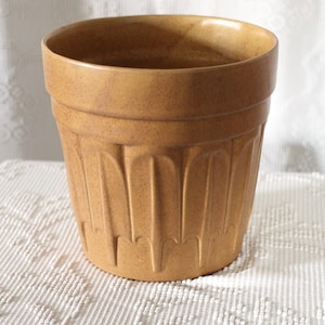 May include: A tan-colored, ceramic plant pot with a textured, ridged exterior. The pot has a wide opening and a slightly flared shape. The pot is sitting on a white, textured surface.