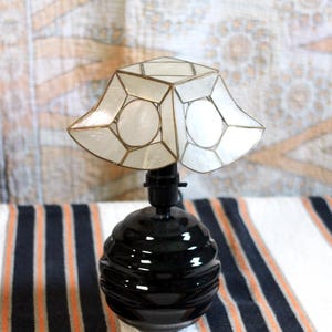 May include: A table lamp featuring a glossy black spherical base with horizontal ridges and a flared, geometric lampshade constructed from translucent white panels held together by thin metallic framing, creating a distinct contrast in shape and color.