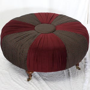 Puede incluir: Un otomán redondo con un diseño de tela marrón y burdeos. El otomán tiene un botón central y está sostenido por cuatro pequeñas patas de madera ornamentadas con ruedas de latón. La forma general es circular.