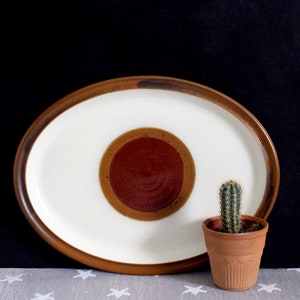 Puede incluir: Un plato de servir ovalado de cerámica blanca con un borde marrón y un diseño circular rojo y marrón en el centro. Una pequeña planta de cactus en una maceta de terracota está sentada sobre el plato.
